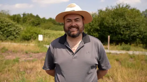 Qays Najm/BBC A man with a polo shirt and a sun hat on standing in a sunny meadow