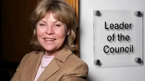 Warwickshire County Council A woman with blonde hair and a brown jacket standing next to a white sign which says Leader of the Council
