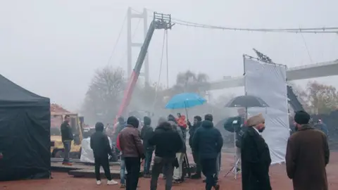 A film crew on location near the Humber Bridge, a towering suspension bridge with a "H"-shaped tower. There are a number of actors in costumes, standing beside crew members and among filming equipment, including tents and cranes.