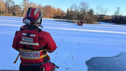 View of an iced over pond, with a light covering of snow. In the foreground is a member of the the fire service, wearing red jacket, marked "rescue" and a helmet and an ear piece. Further along the pond are members of Cumbria fire service. In the background trees are catching the winter sunlight.