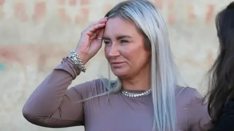 Reuters Lyanne Kennedy falando aos repórteres fora do Tribunal Municipal de Tbilisi. Ela tem longos cabelos loiros e usa joias de prata. Graffiti está na parede atrás dela.