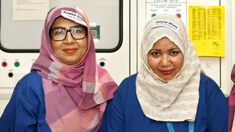 Supplied Ohayla Elhaq and Ayesha Anwar stand close together indoors wearing blue clinical clothing and patterned head coverings. Behind them are wall‑mounted medical system controls, labels, and printed notices.