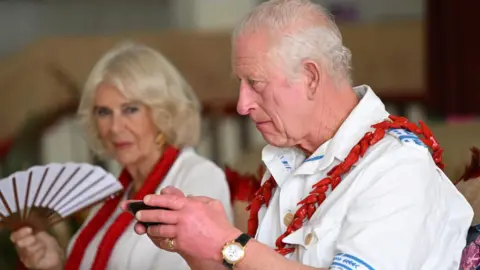 King Charles holding a achromatic  cupful  adjacent  to Queen Camilla