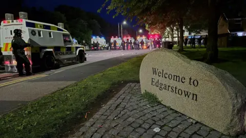 A rock which sayd 'Welcome to Edgarstown' on it on some flagstones with grass and trees surround it. Police land rovers and officers in riot gear can be seen. 