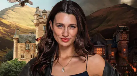 BBC/PA Handout photograph of Elen Wyn, from series three of the Traitors. She wears a diamond necklace with the letter "E" on it and smiles at the camera. Behind her is a UGC image of the Traitors castle. 
