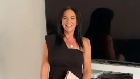 A woman standing in front of a TV and white wall, she is wearing a black dress and has black hair