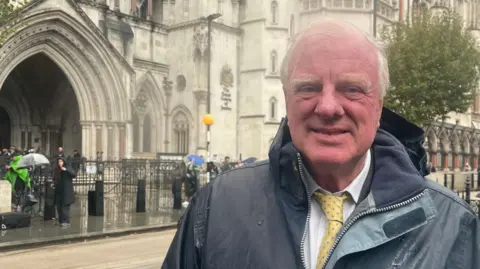 BBC Sir Edward Leigh who has short white hair wearing a black raincoat and white shirt with a yellow tie. It is raining and he is standing in front of the Royal Courts of Justice building.