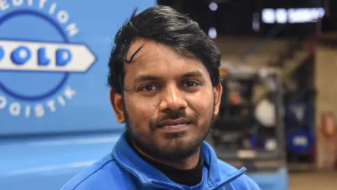 Ajay Kumar Chandapaka standing in front of his blue lorry, and wearing a jumper of the same colour
