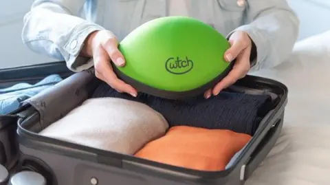 A pair of women's hands hold the green oval Cwtch case over a half packed grey suitcase containing three folded orange, white and black jumpers.