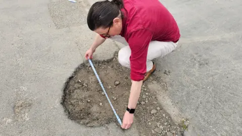 LDRS A man in a red shirt and white jeans crouched down measuring a pothole with a measuring tape