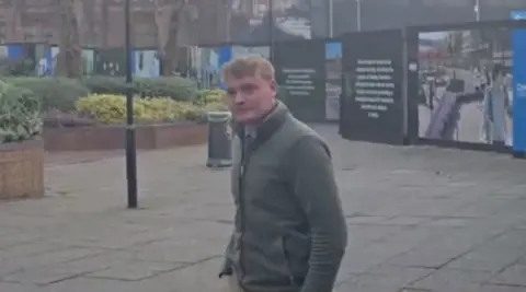 A man with short blonde hair and a grey jacked standing in a paved street with hoardings behind him