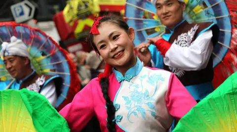 A woman in traditional Chinese dress smiles as she takes part in the parade. She is holding green coloured fans in both hands. 