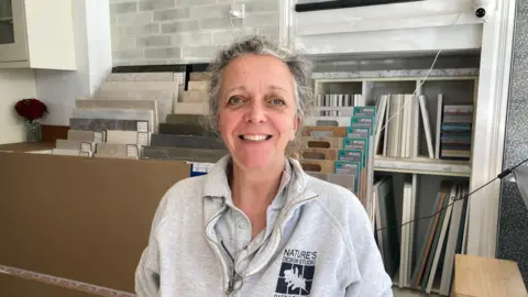 A woman with tied back grey hair smiles while wearing a grey hoodie. She is standing in a shop with samples of flooring behind her.