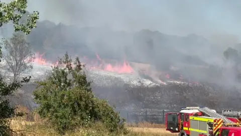 Des Peck Fire at Mawgan Porth on Tuesday. There are fire engines on the right, and fire-damaged land in front. The centre of the land is burning with large plumes of black smoke. 