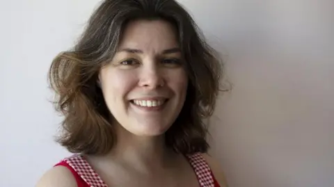 A woman with dark brown hair smiles broadly as she looks into the camera. She is wearing a red and white checked dress.