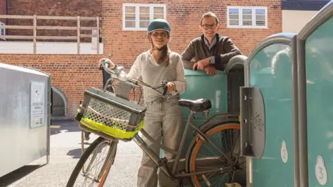 Councillor Hannah Cooper wearing a blue helmet holds a bike with a basket on the front next to three green and grey bike lockers. Chris Jardine, co-founder of Bikedok, is stood behind one of the lockers wearing a brown fleece and round glasses.