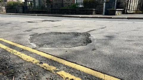 A large circular pothole on a road in Tavistock with a white mark around the outside and next to double yellow lines.
