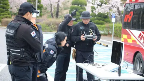 YONHAP/EPA/Shutterstock People in black police uniforms standing by a table set up with a monitor.