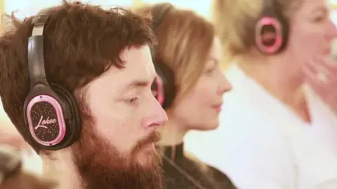 Camsight Alex Innes wearing headphones that are black and pink. He has a dark beard and dark hair. Two other women are to the right of the picture, but they are in the background and slightly blurred. They all have headphones on.