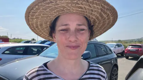 American tourist Katherine Moloney is wearing a black and white striped top and a large straw hat. She has black hair and is standing in a car park