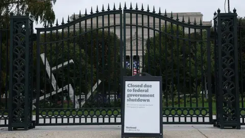 Getty Images Letrero de cerrado afuera de la Galería Nacional de Arte en Washington, D.C. Este letrero está frente a una gran puerta negra de hierro forjado. Al fondo se puede ver un jardín de esculturas y un edificio neoclásico blanco.