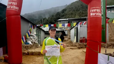 Sally Orange A woman, who is dressed as a Lime, stands at the Everest Marathon finish line. She is holding a medal and a certificate.