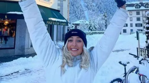 Dyfed Powys Police Katie seen in snow-covered town with arms up and smiling at the camera in a white jacket and black hat.