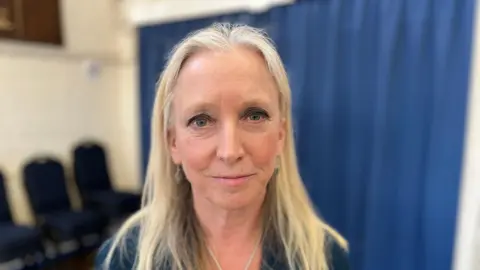 Local MP Roz Savage stands in a community hall with a blue curtain divider in the background.