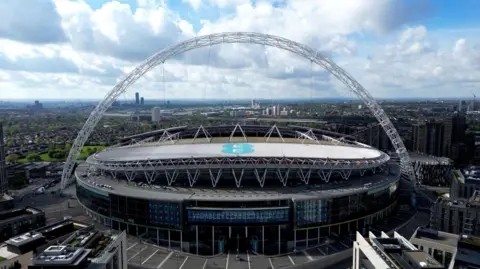 Wembley Stadium from an elevated view, seemingly taken a few hundred metres away. The arch can be seen over the large stadium. There are tower blocks in the distance and white clouds and blue sky above.