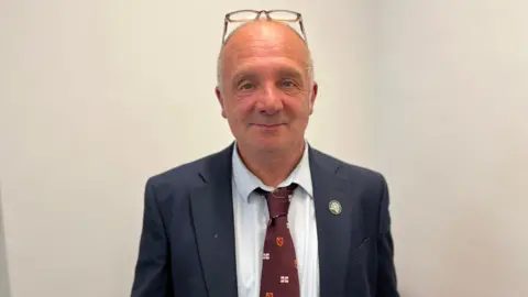 A bald man, with a pair of glasses on his head, looking at the camera with a slight smile. He is wearing a white shirt, a dark blue blazer and a burgundy tie with small Guernsey flags on it.