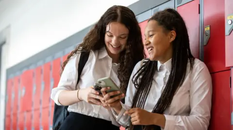 Two teenage girls, dressed successful  achromatic  and achromatic  schoolhouse  uniforms, chatting and looking astatine  their mobile phones successful  the schoolhouse  corridor adjacent  to their lockers (stock image)