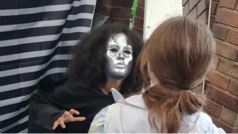 A person, gender unknown, is dressed as "The Unknown" in a black cloak with long black frizzy hair and a silver facemask. A young person in a white shirt with red hair has their back to us and appears to be confronting the Unknown.