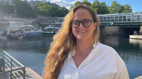 A woman with long wavy strawberry blonde hair in a white shirt and glasses stands in front of a river in the south of France