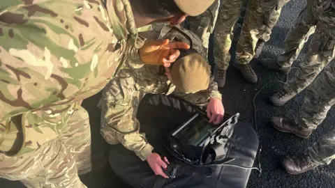 Several soldiers crowd around a small screen on a bag on the ground. Several boots can be seen to the right of the picture