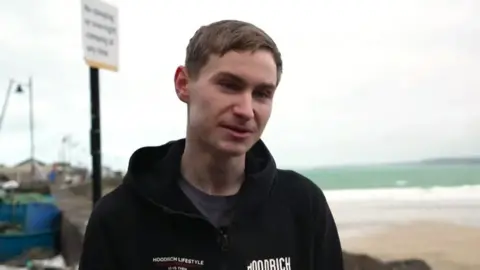 The photo shows a coastal scene with a young person standing on a rocky shoreline. Behind them, the tide is out, exposing a wide stretch of pale sand that curves gently along the bay. The sea looks calm with soft waves rolling in, and the sky is cloudy with a pale, washed‑out light.