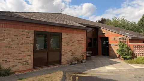 The exterior of a single-storey brick building constructed from light red and brown brickwork with dark brown window frames. The roof is a low-pitched tiled roof extending across the entire structure. Trees and greenery are visible behind and around the building.