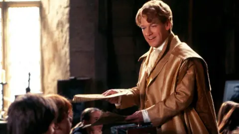 Kenneth Branagh as Gilderoy Lockhart wears a brown jacket and smiles. SINGLE USE