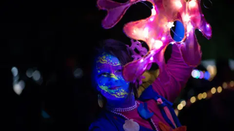 IZZY HENRY A woman in blue paint holding a large object with lights attached