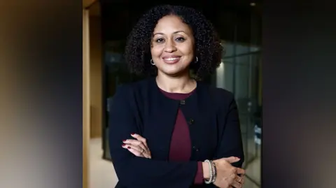 Andrea Simon in a dark suit smiles at camera with arms crossed 