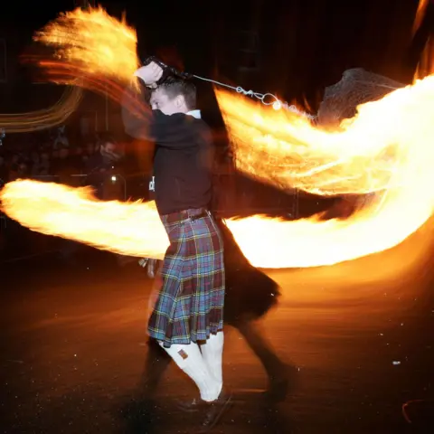 Getty Images A man in a kilt and black shirt with long white socks swinging a flaming fireball around his body at the Stonehaven fireball event 