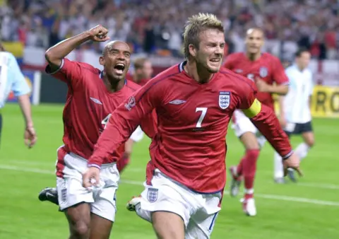 PA Media David Beckham in a red England football shirt runs near the camera, overcome with joy, his arms spread, and behind him is Trevor Sinclair raising his fist in celebration. Other players, including Rio Ferdinand also in a red England shirt, are out of focus in the background on the green football pitch.