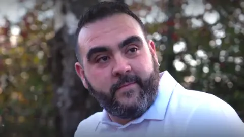 Lee Smith, a man who is sat outside on a bench looking just off to the right of the camera.