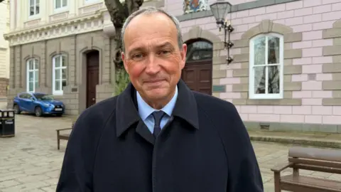 BBC Deputy Lyndon Farnham is wearing a black coat and a blue shirt with a dark blue tie. 