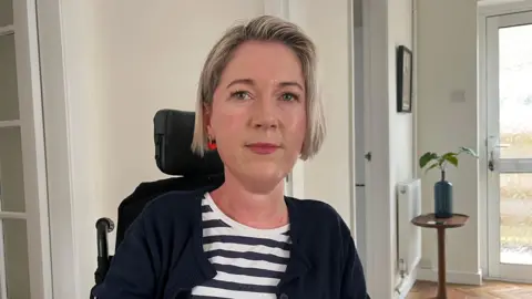 Photo of Sarah, who is looking into the camera and smiling slightly. She has short, chin-length light blonde hair, and is wearing a pair of red earrings. Sarah is wearing a navy blue and white striped top, with a dark blue cardigan over the top. She is pictured in her powered wheelchair, with the back of the chair and the headrest just visible over her shoulder. The photo has been taken indoors, inside her home, which has white walls and a brown wooden floor. On the right of the photo, a glass-panelled door is seen, and next to it is a brown wooden table with a green leafy pot plant on it in a blue vase. 