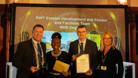 The Royal Wolverhampton NHS Trust Two men and two women, one of whom is Baroness Floella Benjamin, stand in a line and smile a the camera. The man on the far left holds a plaque, Floella Benjamin holds a gold envelope, and another man holds a certificate with the word 'winner' on it.