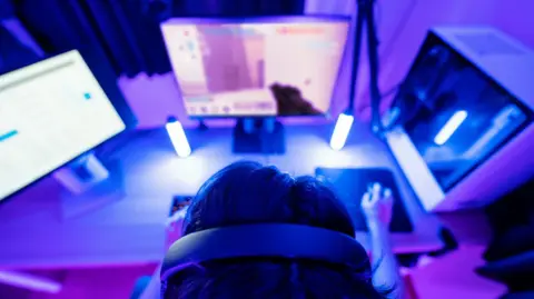 Getty A person is playing a first-person shooter video game at a monitor. 
They have a large headset on and their hand is on a mouse.
A number of UV lights are visible on their table where the monitor and gaming system is located. 