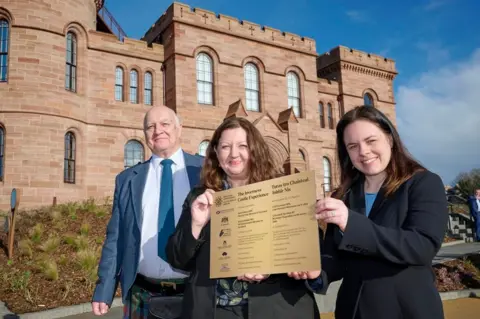 Highland Council Three people stand in front of Inverness Castle they are, from left to right, a man in a kilt with a blue jacket and tie and a bald head, a woman with long reddish-brown hair and a woman with dark hair and a dark jacket. The two women are holding a golden sign describing the castle experience.