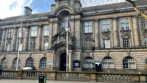 The council building is made up of large grey, stone blocks and paned windows. There are flags at the entrance and a low wall runs around the front of the building.