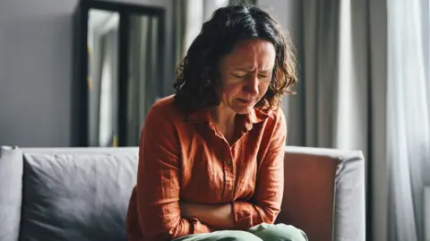 Stock image of woman with curly brown shoulder length hair sitting on a grey softa. Her arms are folded over her abdomen and she appears to be grimacing and upset in pain. She wears a terracotta shirt and green trousers. There is light coming in through a nearby window. 