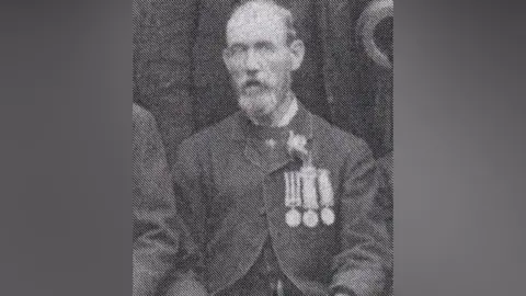 Noonans The image is a black-and-white, grainy historical photograph showings a seated man facing the camera and seated upright, wearing a dark suit, with several medals pinned to the chest. 